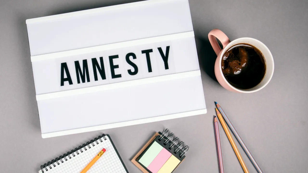 Lightbox sign reading 'AMNESTY' with a coffee mug, pencils, notebook and sticky notes on a grey desk.